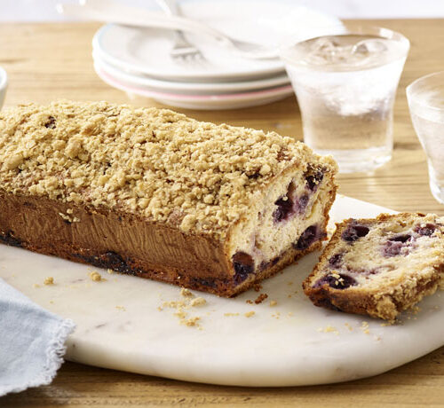 A loaf of blueberry crumb cake with one slice cut, served on a marble board with two glasses of water and plates.