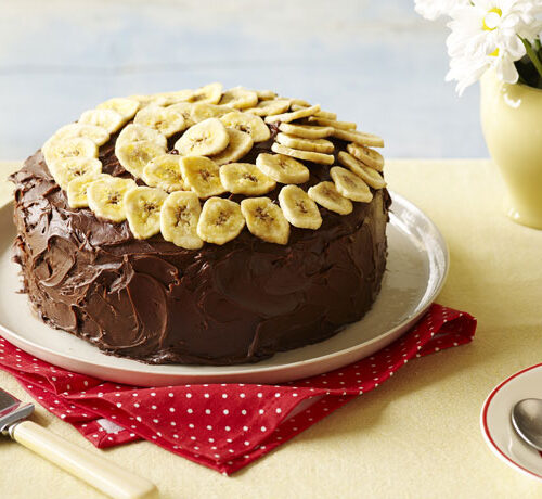 A chocolate frosted cake topped with banana slices on a plate, next to a cup of coffee and a vase of daisies.