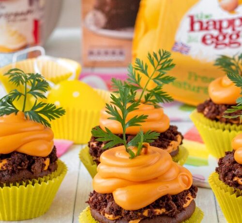 Chocolate cupcakes with orange frosting and carrot tops, arranged on a table with baking ingredients in the background.