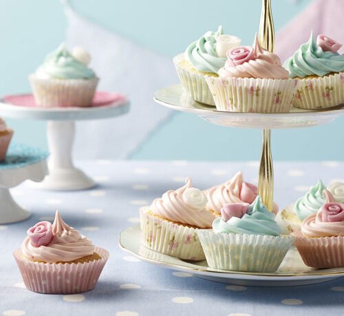 Cupcakes with pastel frosting and rose decorations arranged on cake stands and a polka dot tablecloth.