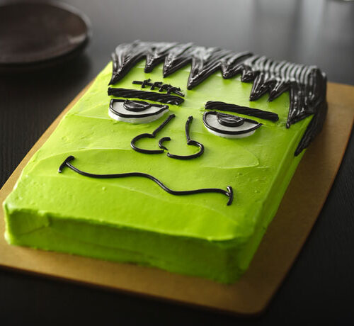 Rectangular cake decorated as Frankenstein’s monster with green icing and black frosting details on a brown board.