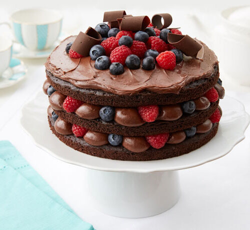 Three-layer chocolate cake with berries and chocolate curls on top, displayed on a white cake stand.