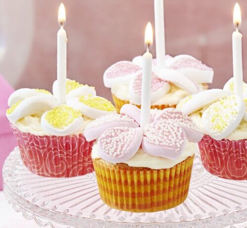 Four decorated cupcakes with lit candles on a glass stand, topped with white icing and pastel candy pieces.