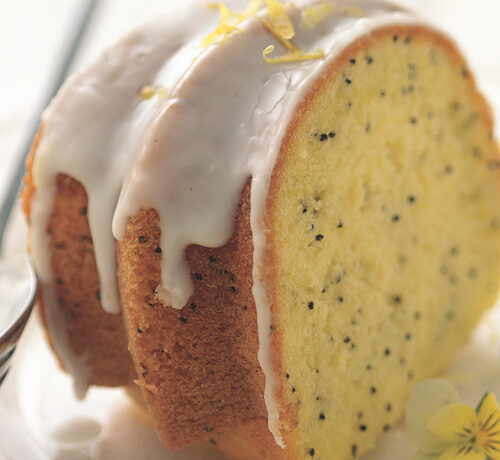 A slice of lemon poppy seed Bundt cake with white icing on a plate, garnished with lemon zest and a flower.