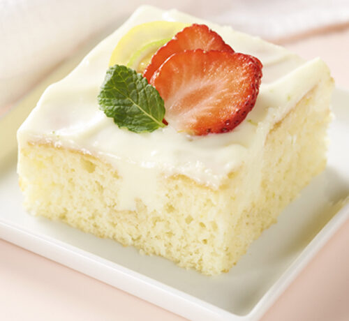 A slice of frosted yellow cake topped with fresh strawberry slices, a lemon wedge, and a mint leaf on a white plate.