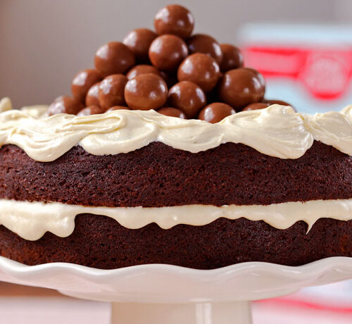 A chocolate cake with white frosting and chocolate balls piled on top, displayed on a white cake stand.