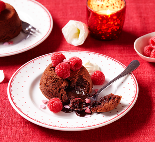 Chocolate lava cake topped with raspberries on a white plate, with a fork and extra raspberries on the side.