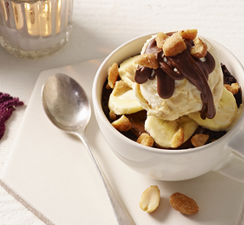 A cup with ice cream, banana slices, nuts, and chocolate syrup, next to a spoon and a purple napkin.