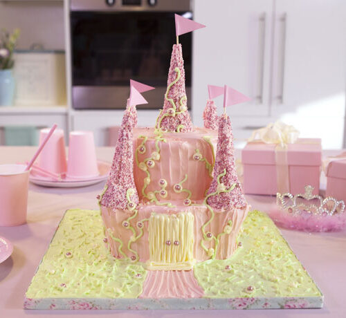 A pink castle-shaped birthday cake with flags, surrounded by pink party supplies and gift boxes on a table.