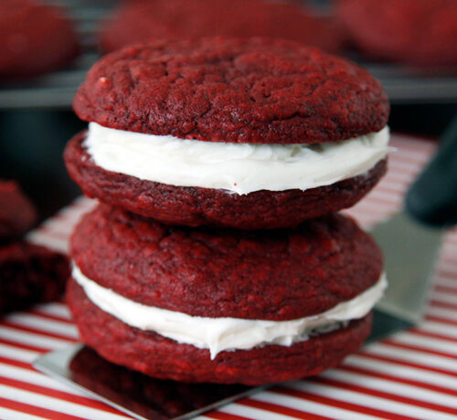 Two red velvet whoopie pies with white cream filling are stacked on a striped surface, with more in the background.