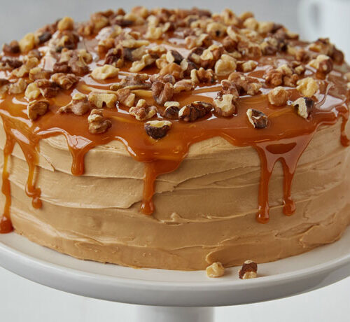 Round layer cake with caramel frosting, caramel drizzle, and chopped walnuts on top, displayed on a white cake stand.