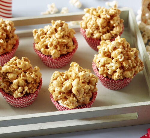 Six popcorn balls in red cupcake liners on a white tray, with popcorn and drinks in the background.