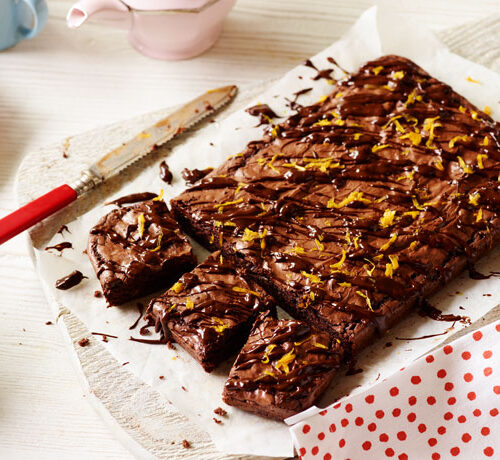 A tray of chocolate brownies topped with orange zest, partially sliced, on parchment paper with a knife beside it.
