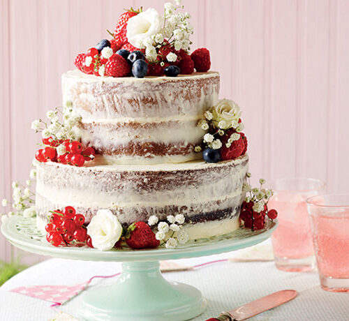 Two-tier naked cake topped with berries and white flowers on a green stand, next to glasses of pink drink.