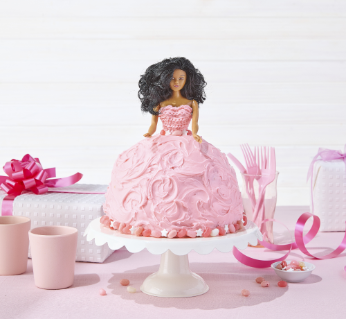 A doll cake with pink frosting on a white stand, surrounded by pink tableware, gifts, and decorations.