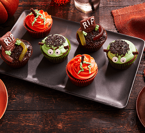 A tray of Halloween-themed cupcakes decorated as monsters and graves, with orange and green frosting.
