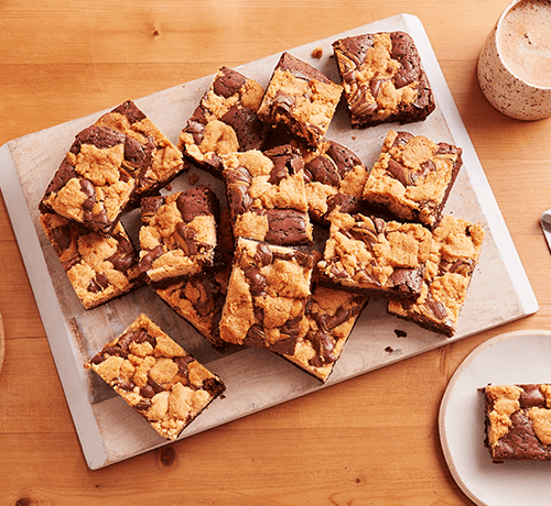 peanut butter and chocolate cookie bars on a white platter surrounded by two mugs and two small plates with 1 cookie bar on each