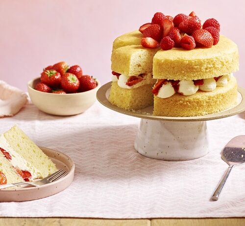 A two-layer sponge cake with cream and strawberries on a stand, with a slice served on a plate nearby.