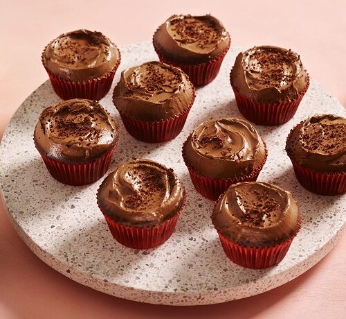 Nine chocolate-frosted cupcakes on a round tray, with two cups of coffee and an empty plate nearby.