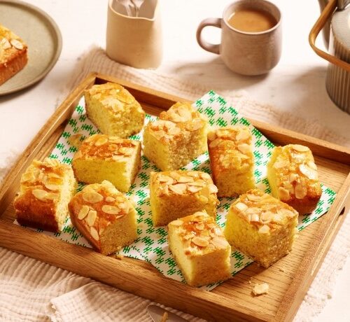 A wooden tray with almond-topped cake squares, two cups of tea, a teapot, and plates on a light table.
