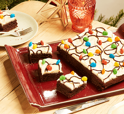 Rectangular chocolate cake with white icing, chocolate drizzle, and colorful candies, cut into square pieces.