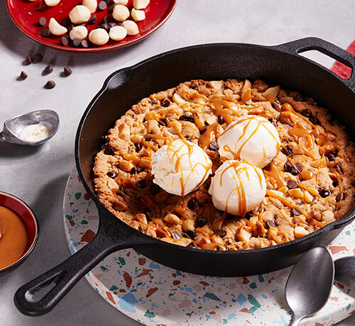 A skillet cookie topped with two scoops of vanilla ice cream and caramel drizzle, surrounded by bowls and spoons.