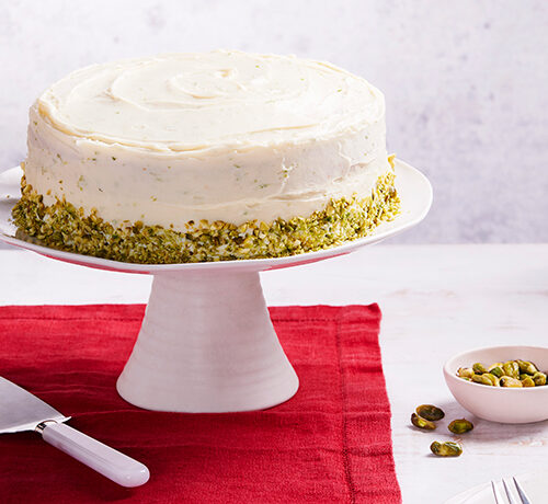 A frosted cake with chopped pistachios on a white stand, next to a teapot, cup, and limes on a red cloth.