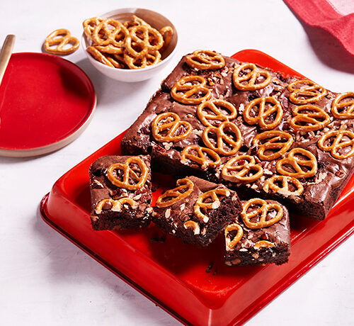 A tray of chocolate brownies topped with pretzels, with coffee, caramel pieces, and pecans nearby on a white table.